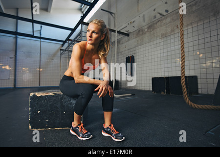 Young woman sitting on a box at gym after her workout. Caucasian female athlete taking rest after exercising at gym. Stock Photo