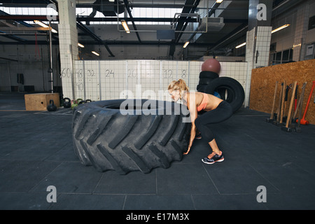 Tough female athlete flipping a huge tire. Young woman doing crossfit exercise at gym. Stock Photo