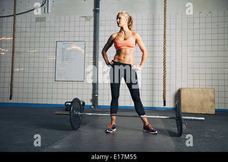 Full length portrait of muscular young woman standing at gym looking away with barbells on floor. Strong crossfit female at gym. Stock Photo