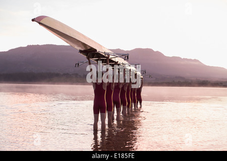 Rowing team carrying row boat overhead in still lake Stock Photo