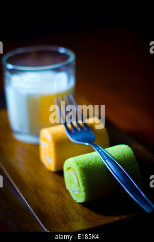 Two “Swiss rolls” slices green orange yellow on a tray have a fork on top with fresh Plain milk in glass Stock Photo