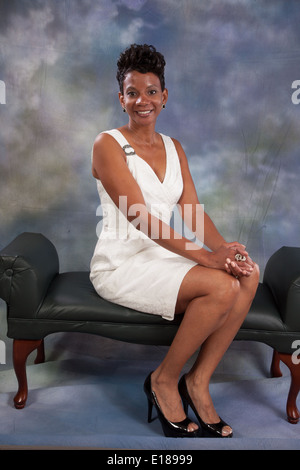 Pretty black woman in white dress, sitting on a bench with her hands on her knees, smiling at the camera Stock Photo