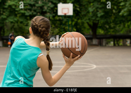 Young slender teenage girl playing basketball throwing the ball, view from behind Stock Photo