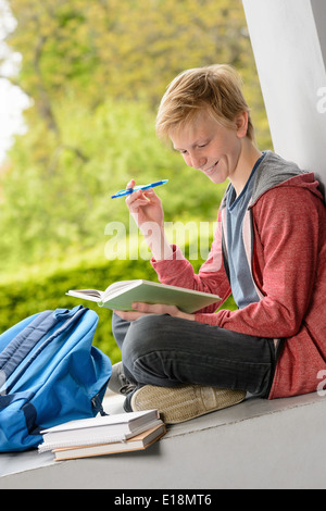 Smiling teenage boy reading book sitting on wall outside school Stock Photo