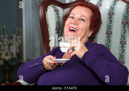 Senior Woman having good time while having her Turkish Coffee, she is wearing a purple sweater and sitting on comfortable seat Stock Photo