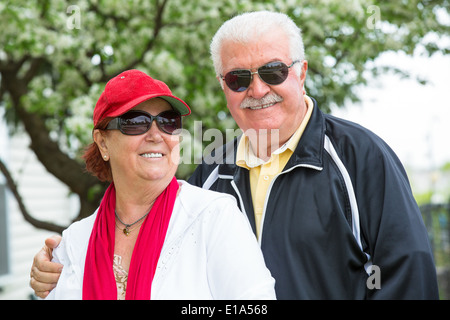 Easygoing positive senior couple posing with their sports clothing and sunglasses in front of blooming spring tree. Man holding Stock Photo