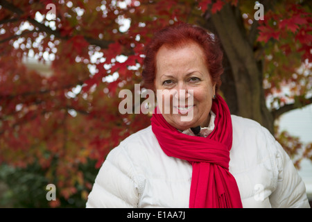 Red hair senior lady looking at you happily and trustfully with her red scarf and white coat under the tree with red falling lea Stock Photo