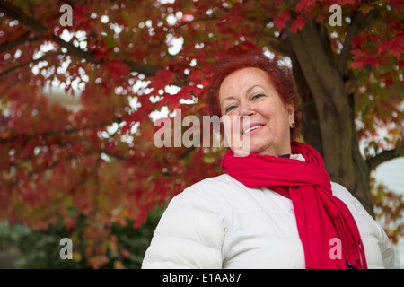 Red hair senior lady under the tree with red leaves looking at you happily with her red scarf and white coat Stock Photo