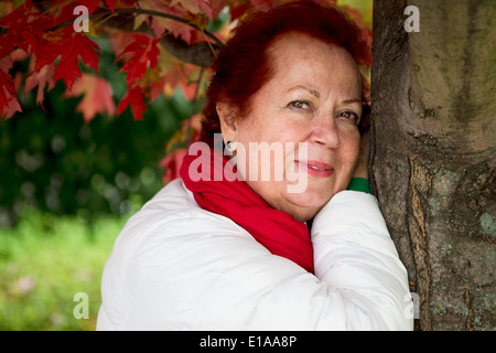 Red hair senior lady looking at you thoughtfully with her red scarf and white coat while leaning to the tree Stock Photo