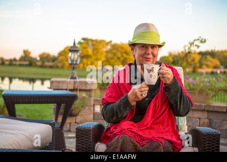 Senior woman with a straw green hat, sitting at the patio and showing her hot tea, perhaps she wants to stay warm Stock Photo