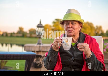 Senior woman with a straw green hat, sitting at the patio and blowing her hot tea, perhaps she wants to stay warm Stock Photo