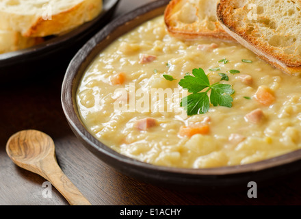 A rustic bowl of hearty spit pea soup with smoked ham, carrots, potato, and french bread. Stock Photo