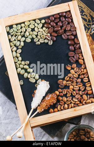 Green, brown unroasted decaf and black coffee beans with sugar sticks on black empty chalkboard over vintage books. Top view. Stock Photo