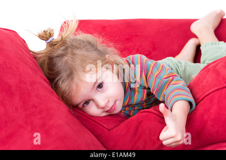 Beautiful little girl giving a thumbs up gesture as she lies comfortably on her stomach on a red couch looking at the camera wit Stock Photo