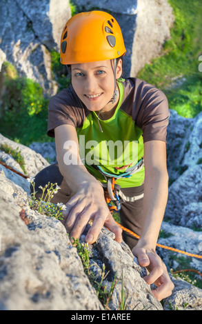 Smiling woman, rock climber in yellow helmet reaching top of mountain during sunset. Stock Photo