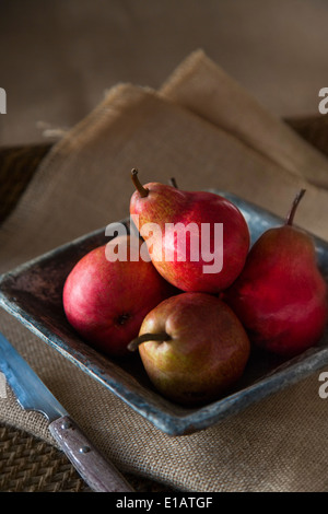 group of red pears in rustic ceramic dish, with knife, on sack cloth and woven tray. Stock Photo