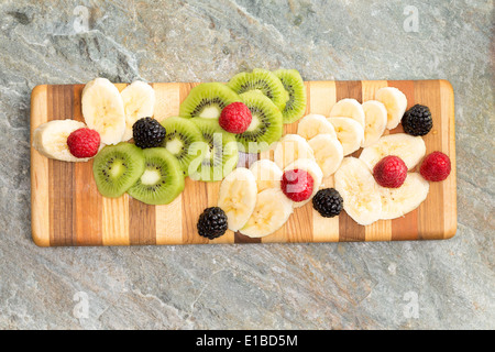 Sliced fresh ingredients ready for a fruit salad displayed on a decorative wooden striped chopping board including tropical kiwi Stock Photo