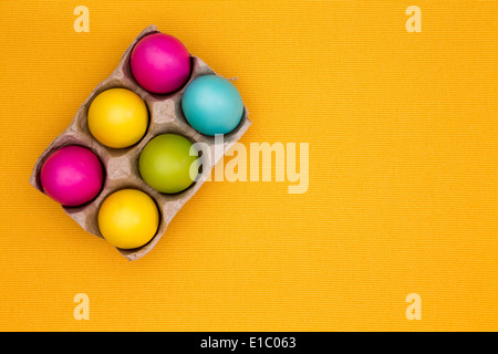 High-angle close-up of six vivid colored Easter eggs in a carton, on a table covered by a yellow cotton tablecloth Stock Photo