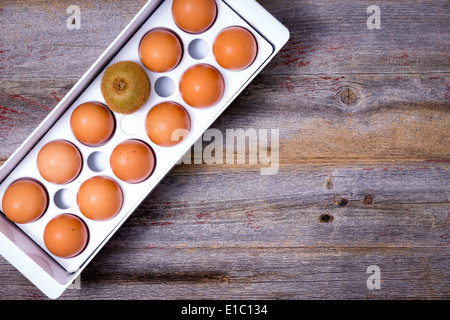 View from above of a single different kiwifruit stored in a tray of farm fresh brown eggs on an old weathered wooden kitchen cou Stock Photo