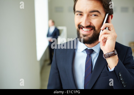 Image of young businessman speaking on the phone and looking at camera on background of his colleague Stock Photo
