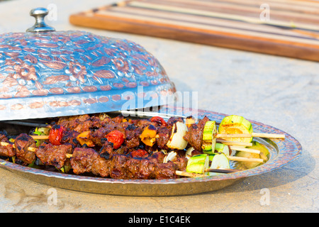 Tasty grilled beef and vegetable kebabs on metal plate with open domed lid on concrete counter in an outdoor summer kitchen Stock Photo