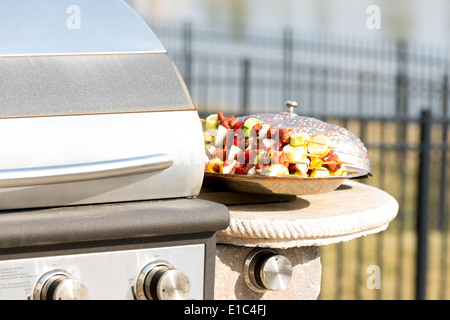 Delicious gourmet kebabs ready to be barbecued standing on a metal plate in the summer sunshine alongside a closed gas BBQ Stock Photo