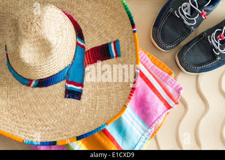 Wide brimmed straw sombrero lying on a colorful striped beach towel with a pair of sneakers on tropical beach sand with a decora Stock Photo