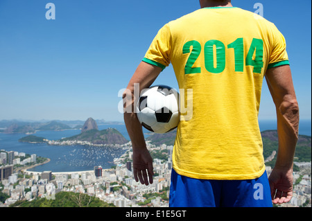 Brazilian football player in 2014 shirt standing with football bright sunny Rio de Janeiro skyline with Sugarloaf Mountain Stock Photo