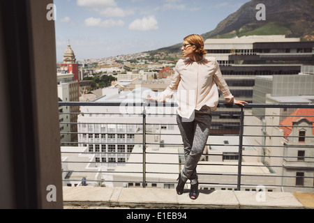 Beautiful young woman standing on a balcony looking away with a cup of coffee. Caucasian female model posing on a terrace. Stock Photo