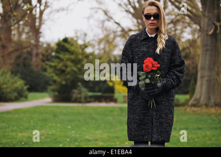 Portrait of sad young woman in black dress at cemetery holding fresh roses. Female at graveyard with flowers. Stock Photo