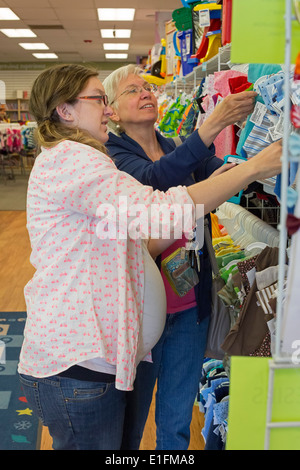 A pregnant 26-year-old woman shops for baby clothes at a resale shop, helped by her mother. Stock Photo