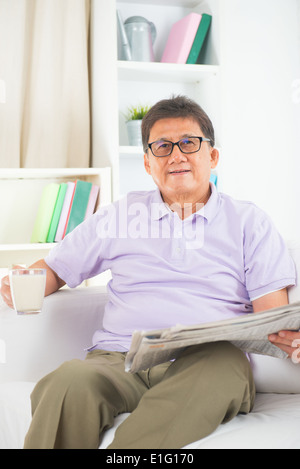 senior asian chinese reading newspaper and drinking milk on a sofa in home Stock Photo