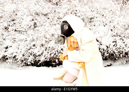Woman dressed in white wearing a gas mask and hugging a teddy bear during snow fall Stock Photo