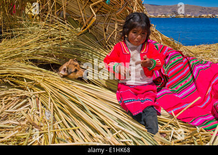 Young girl of the Uro Indians, about 5 years old, wearing traditional dress, sitting next to a dog on reed bundles, floating Stock Photo