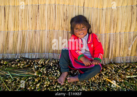 Smiling young girl the Uro Indians, about 6 years old, wearing traditional dress, sitting in front of a reed hut, floating Stock Photo