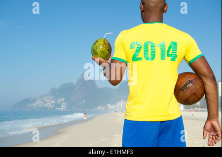 Brazilian football player wearing 2014 shirt in Brazil colors holding vintage soccer ball drinking coconut Rio de Janeiro Stock Photo