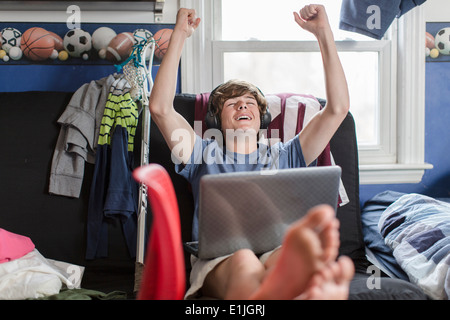 Teenage boy sitting on chair using laptop computer, arms raised Stock Photo
