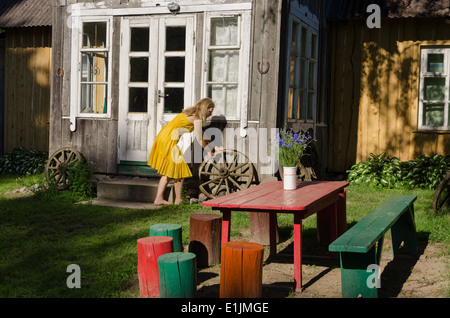 Rural wooden house yard and woman girl in yellow dress rolls old carriage wheel. Beautiful retro decoration. Colorful chairs and Stock Photo