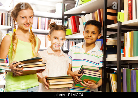 Three friends with books standing near bookshelf Stock Photo