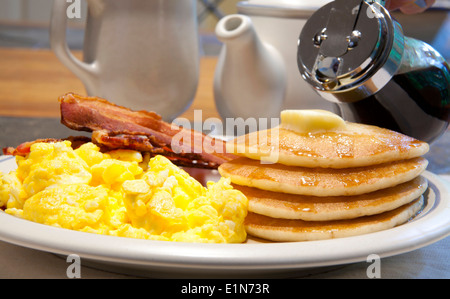 Breakfast of pancakes, scrambled eggs and bacon on a white plate being covered in maple syrup Stock Photo