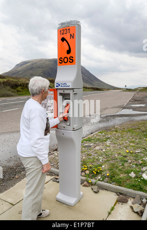 Senior woman using an emergency SOS telephone beside a remote country road calling for roadside assistance help. Glen Coe Highland Scotland UK Britain Stock Photo