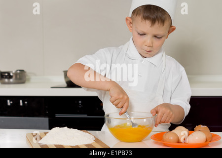 Young boy in a chefs toque and apron standing at a kitchen counter whisking eggs with a fork as he learns to bake Stock Photo