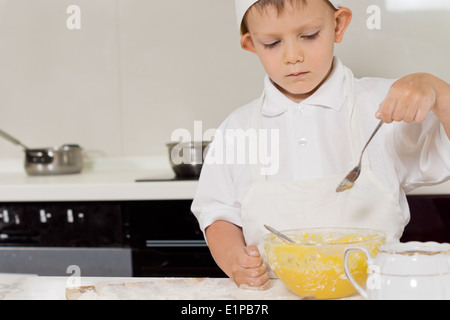 Little boy in a chefs toque and apron standing at the kitchen counter checking the consistency of the batter in his mixing bowl Stock Photo