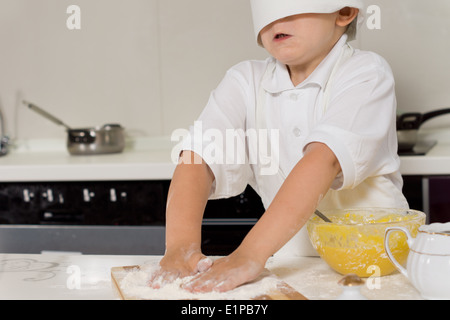 Little child baking in a chefs toque that is falling over his eyes as he rubs his hands in the flour while making pastry Stock Photo