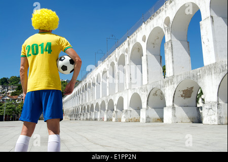 Brazilian football player with yellow afro wig holding soccer ball wearing 2014 shirt in Brazil colors Rio de Janeiro Stock Photo