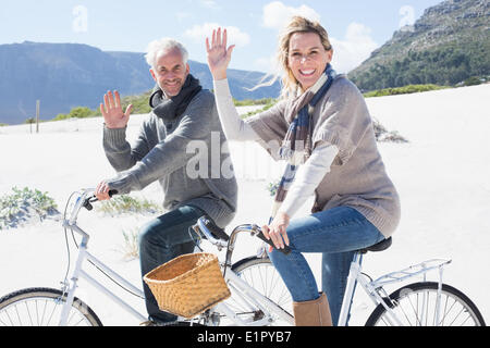 Carefree couple going on a bike ride on the beach waving at camera Stock Photo