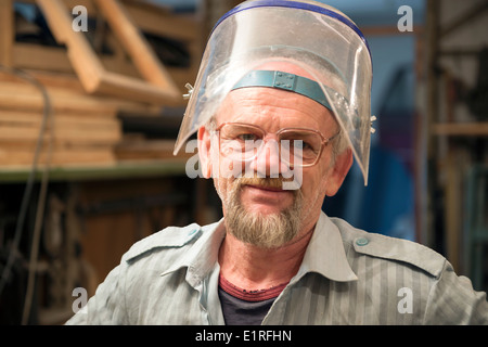 The aged man stands inside his carpentry workshop with the face shield on his head. Stock Photo