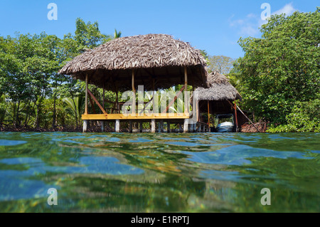 Tropical hut with thatch roof overwater and a boathouse with vegetation in background, viewed from water surface, Caribbean sea Stock Photo