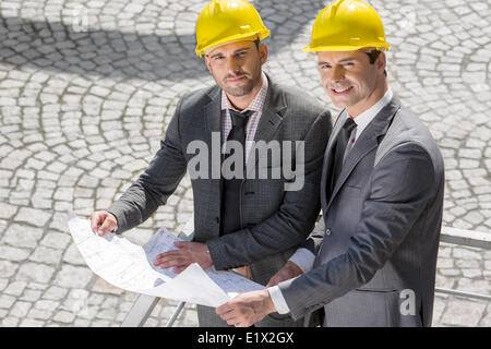 Portrait of confident young businessmen in hard hats examining blueprint outdoors Stock Photo