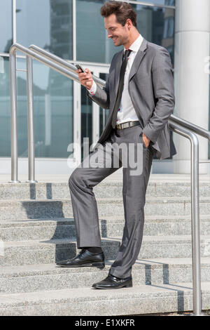 Full length of smiling businessman text messaging through cell phone while standing on steps outside office Stock Photo